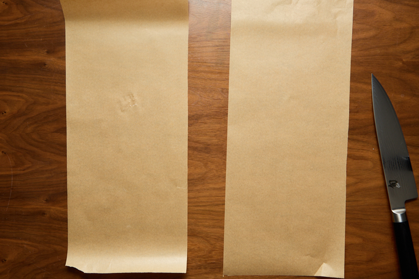 Two rectangular sheets of brown parchment paper are placed side by side on a wooden surface. A large kitchen knife with a black handle lies to the right of the parchment sheets.