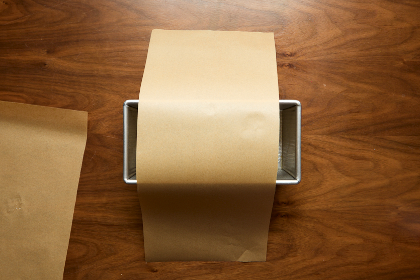 A metal loaf pan sits on a wooden surface with a sheet of brown parchment paper placed inside, extending over two sides of the pan. Another sheet of parchment is partially visible to the left.