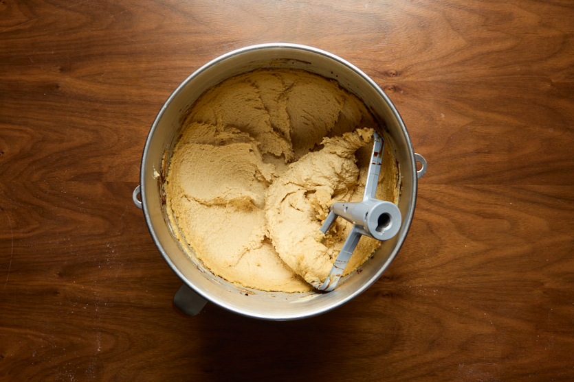 A metal mixing bowl with pale yellow cookie dough and a paddle attachment sits on a wooden surface. The dough appears partially mixed, with the paddle resting in the bowl.