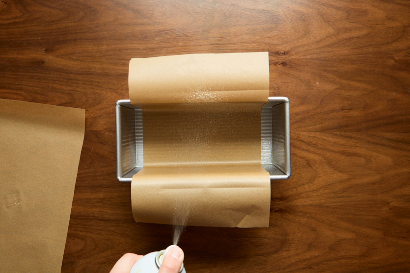 A rectangular metal baking pan lined with parchment paper is being sprayed with non-stick cooking spray. The scene is set on a wooden surface.