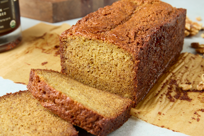 A loaf of banana bread sits on brown parchment paper with two slices cut in front. The bread has a golden-brown crust and a moist, dense texture. Some walnuts are visible in the background.