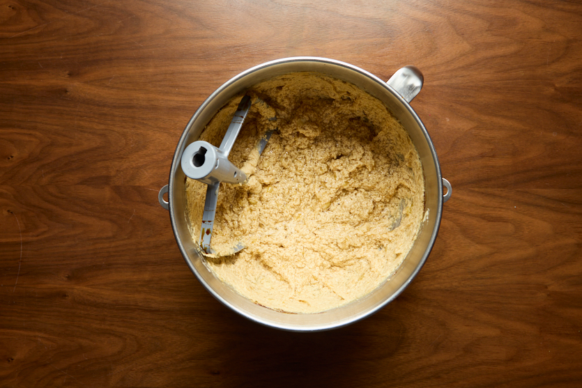 A metal mixing bowl with pale yellow, thick cookie dough and a paddle attachment sits on a wooden surface.
