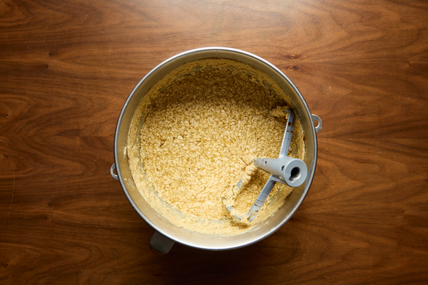 A metal mixing bowl containing a pale, coarse batter with a paddle attachment sits on a wooden surface, viewed from above.