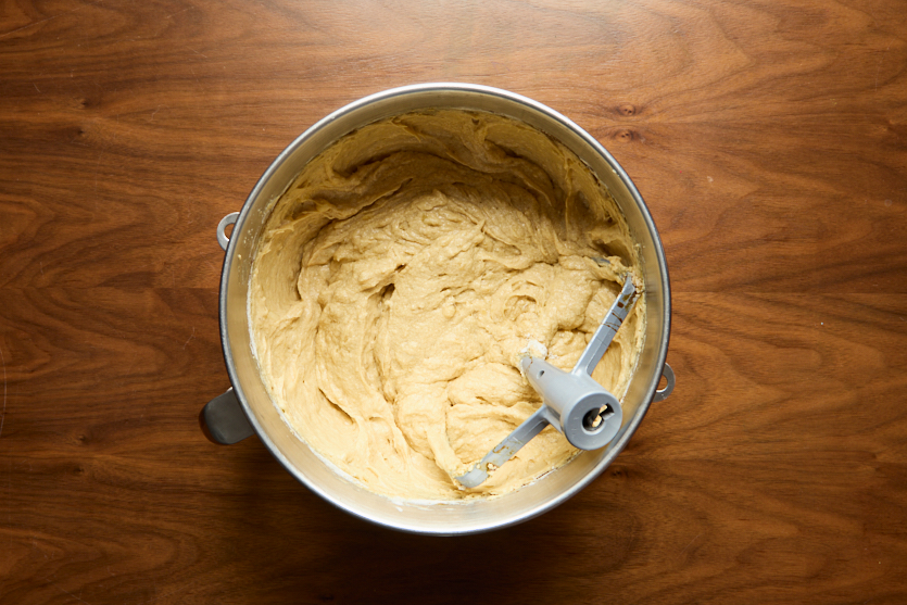 A metal mixing bowl filled with creamy, light-brown dough sits on a wooden surface. A paddle attachment is partially buried in the dough, suggesting it was recently mixed.