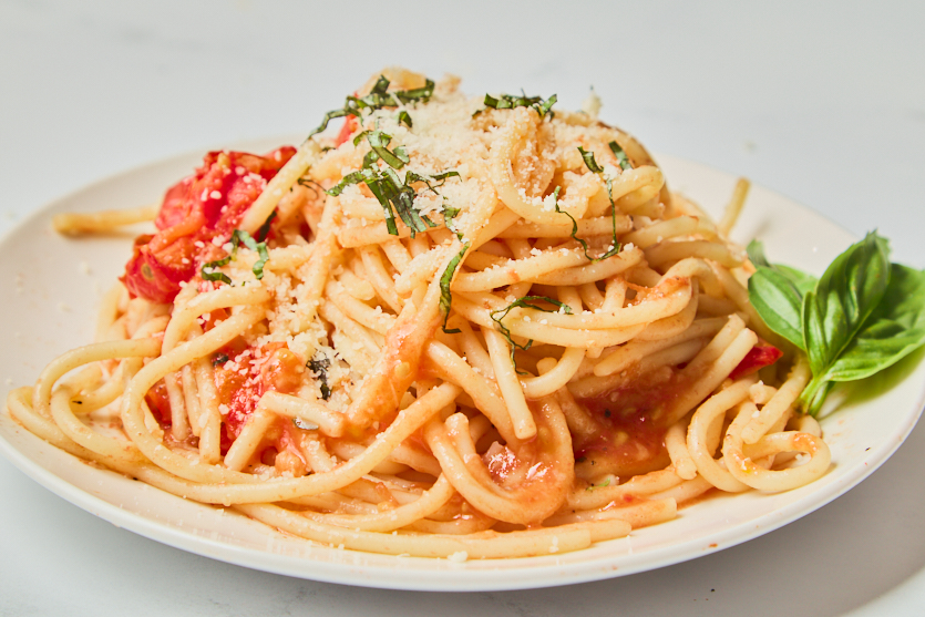 A plate of spaghetti with tomato sauce, grated cheese, and chopped herbs, garnished with a sprig of fresh basil on the side, sits on a white surface—an inviting display of classic Italian food reminiscent of traditional Pasta al Pomodoro.